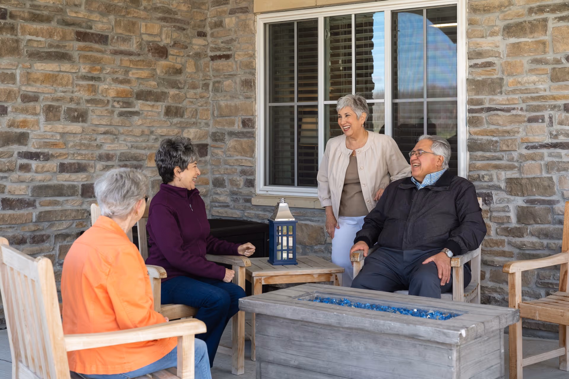 Four elderly people sitting and standing around a wooden outdoor fire pit table on a patio with stone walls and a window in the background, smiling and engaging in conversation.