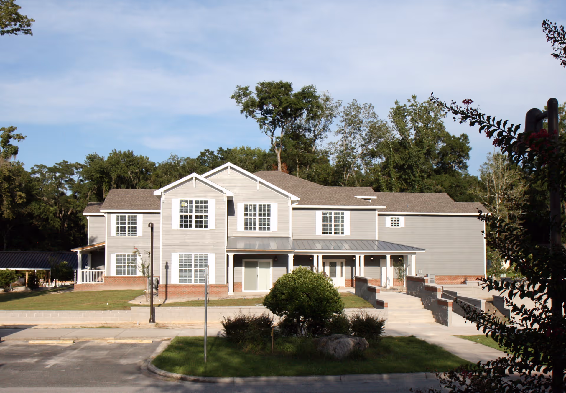 Exterior view of a two-story residential building with gray siding and white trim, surrounded by greenery and trees under a partly cloudy sky. There is a small landscaped area with bushes and a rock in front of the building, along with a paved driveway and parking spaces.