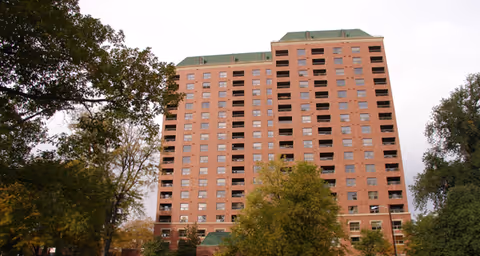 Front view of a tall brick multi-story residential building surrounded by trees under an overcast sky.