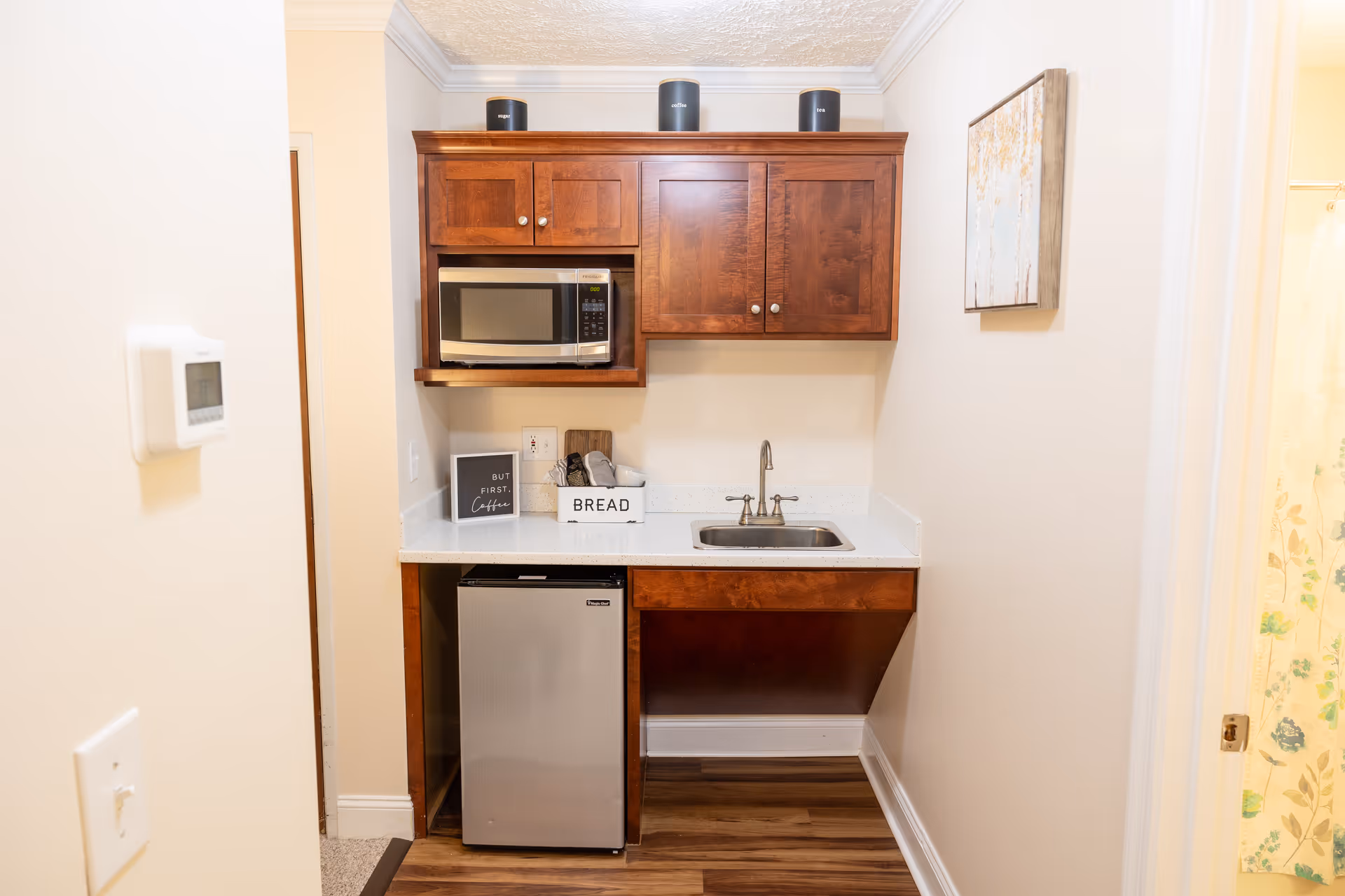 A small kitchenette with wooden cabinets, a microwave, a mini refrigerator, a sink, and a countertop. On the countertop are containers labeled sugar, coffee, and tea, a bread box, and a small sign that says 'BUT FIRST, Coffee'. There is a piece of wall art on the right wall and a thermostat on the left wall.