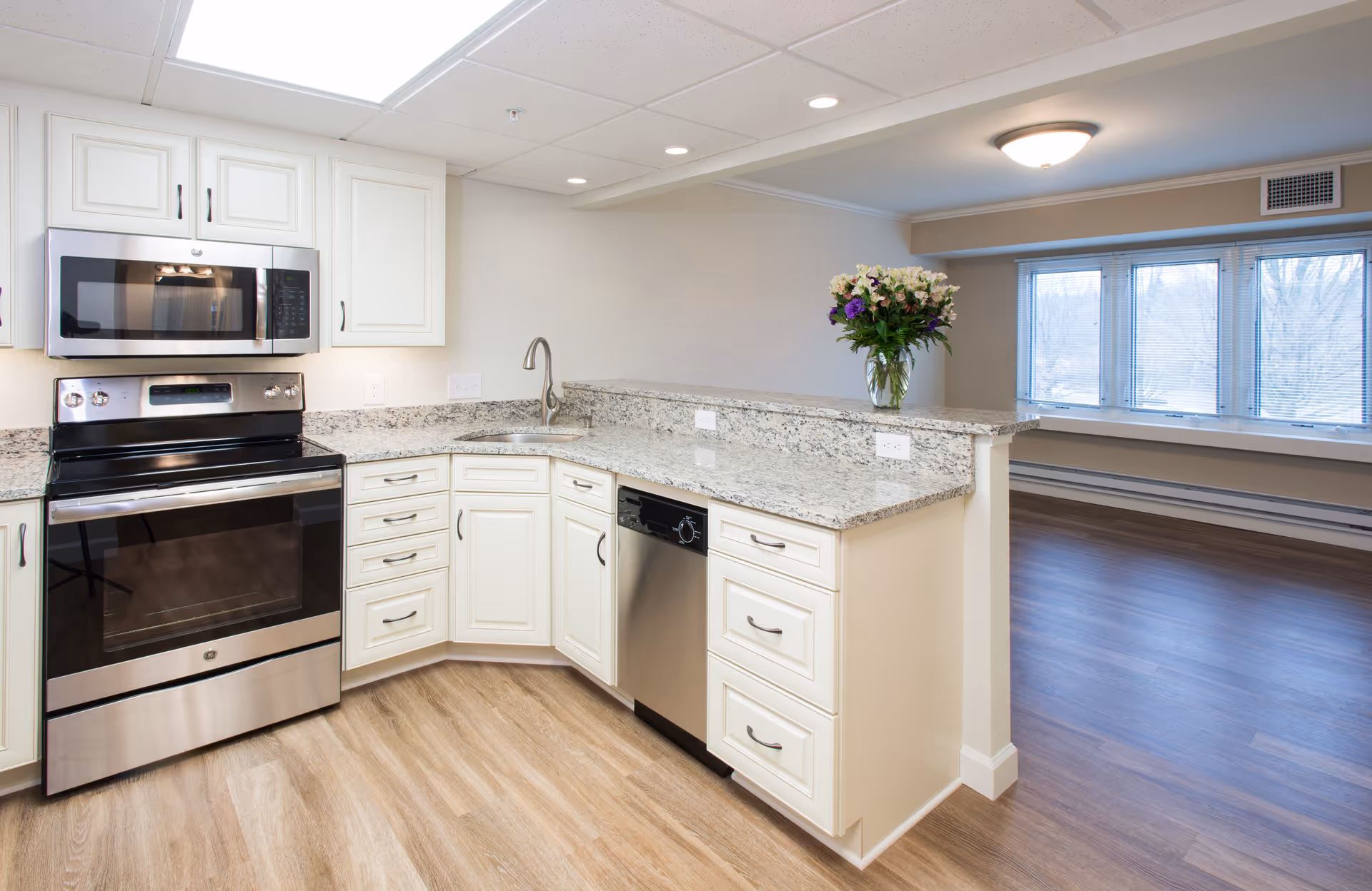 Modern kitchen with white cabinets, granite countertops, stainless steel appliances including a microwave, stove, and dishwasher. There is a vase with flowers on the counter and a large window in the adjacent room letting in natural light.