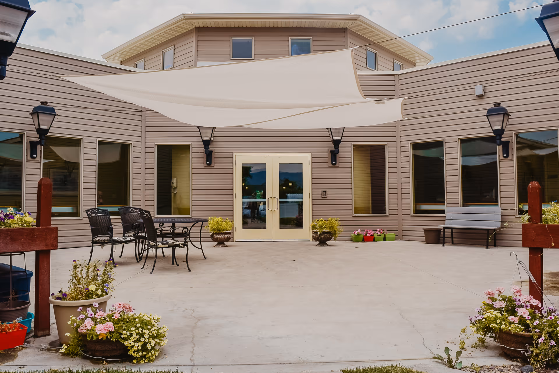 Outdoor courtyard area of a senior living facility with beige siding walls, several windows, double glass doors, a white shade sail overhead, black metal chairs and tables, benches, potted plants, and decorative street lamps.