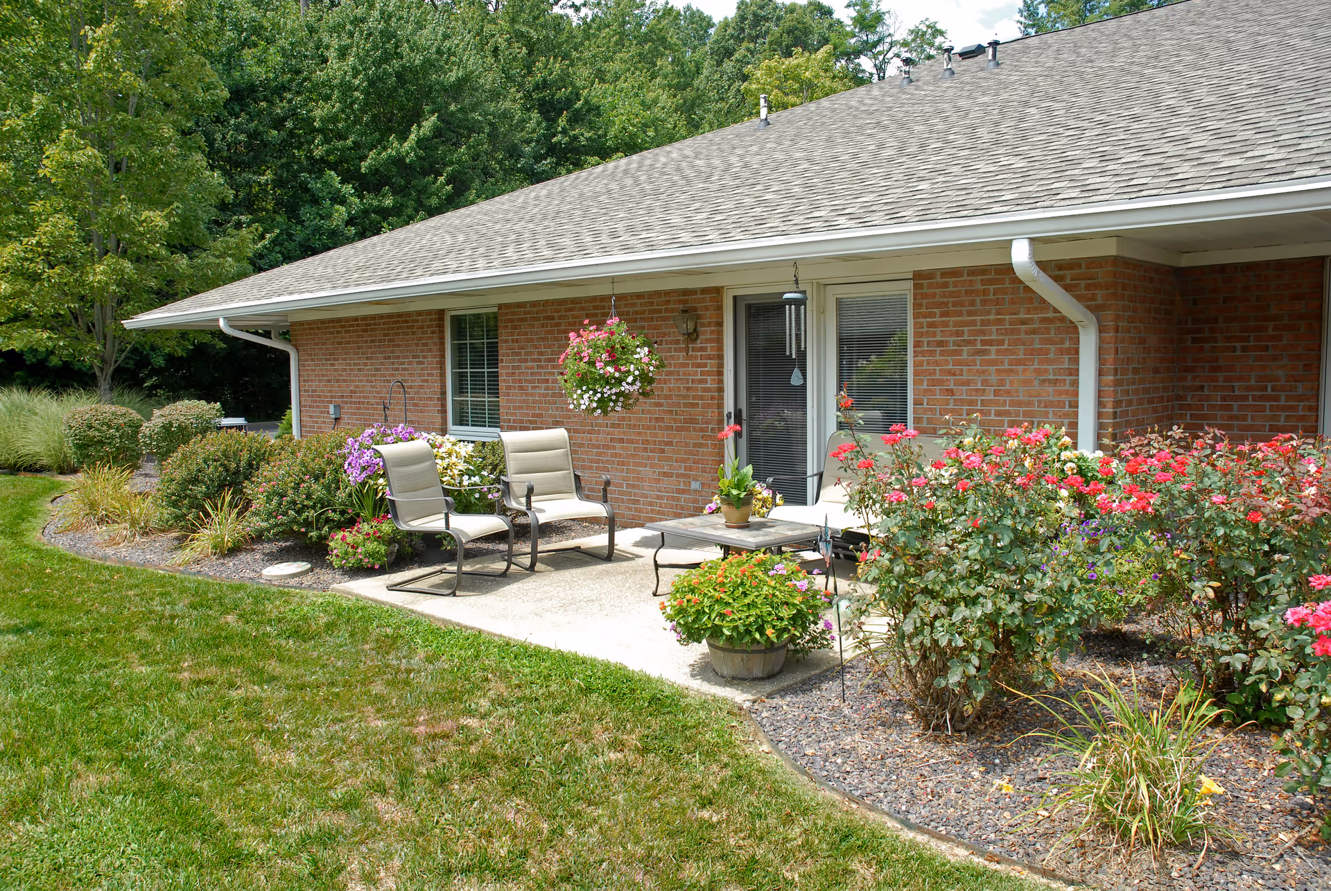 A brick single-story building with a small patio holding two chairs, a table, hanging and potted flowers, and a landscaped garden.