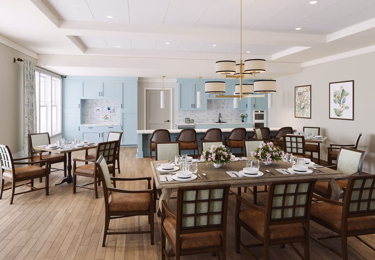A bright dining room with wooden floors and multiple tables set for meals. The room features a large wooden dining table in the foreground with chairs around it, decorated with flower arrangements and place settings. In the background, there is a light blue kitchen area with cabinets, a countertop, and bar stools. The walls are adorned with framed botanical artwork, and large windows with patterned curtains allow natural light to fill the space.