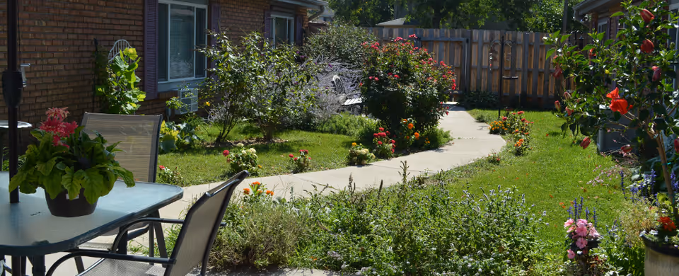 A sunny outdoor garden area with a curved concrete pathway surrounded by green grass, colorful flowers, and shrubs. There is a glass-top table with two chairs and a potted plant on the table. A brick building with windows is visible on the left side, and a wooden fence encloses the garden in the background.