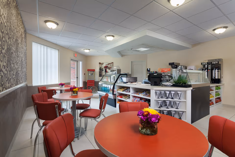 Bright café/dining area with red tables and chairs, a service counter with display cases and coffee machines, and small floral centerpieces.
