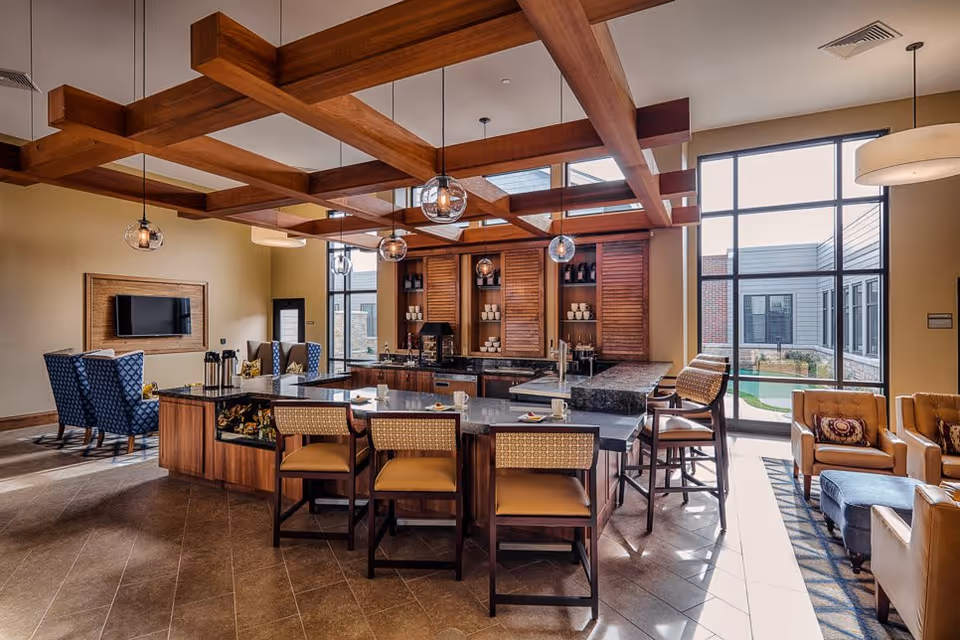 A spacious, well-lit common area with a large kitchen island featuring bar stools, pendant lights hanging from a wooden beam ceiling, and a seating area with armchairs and a TV mounted on the wall. Large windows allow natural light to fill the room and offer a view of an outdoor courtyard.