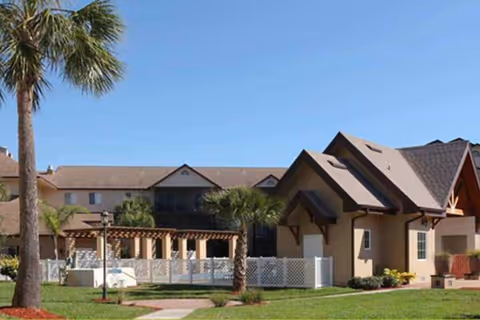 Exterior view of a senior living facility with a beige building featuring a peaked roof, surrounded by palm trees, green grass, and a white fence under a clear blue sky.