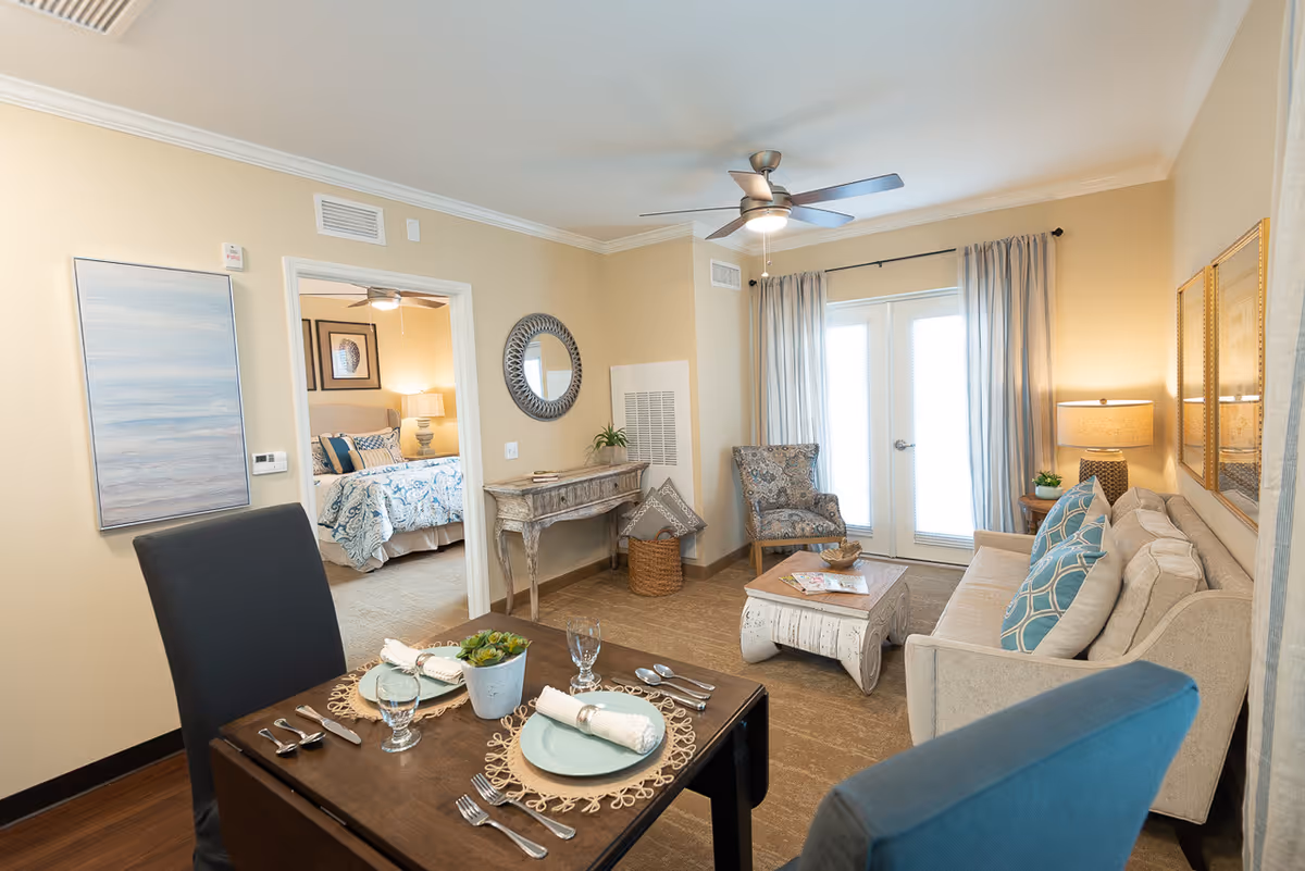 A cozy living and dining area in a senior living facility with a small dining table set for two, a beige sofa with blue patterned pillows, an armchair, a coffee table, and a console table with a round mirror above it. The room has light beige walls, a ceiling fan, and French doors with curtains leading outside. A bedroom with a bed and bedside lamp is visible through an open doorway.