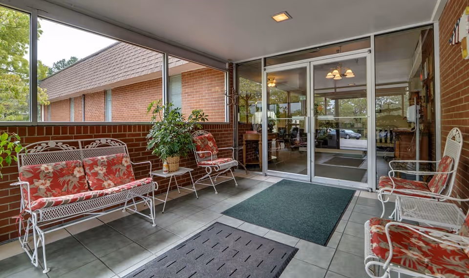 Enclosed porch area with white metal framed chairs and a bench featuring red floral cushions. There are two green floor mats on tiled flooring, potted plants on small tables, and large windows showing the outside greenery and a brick building. Glass double doors lead into an interior space with visible ceiling lights and furniture.