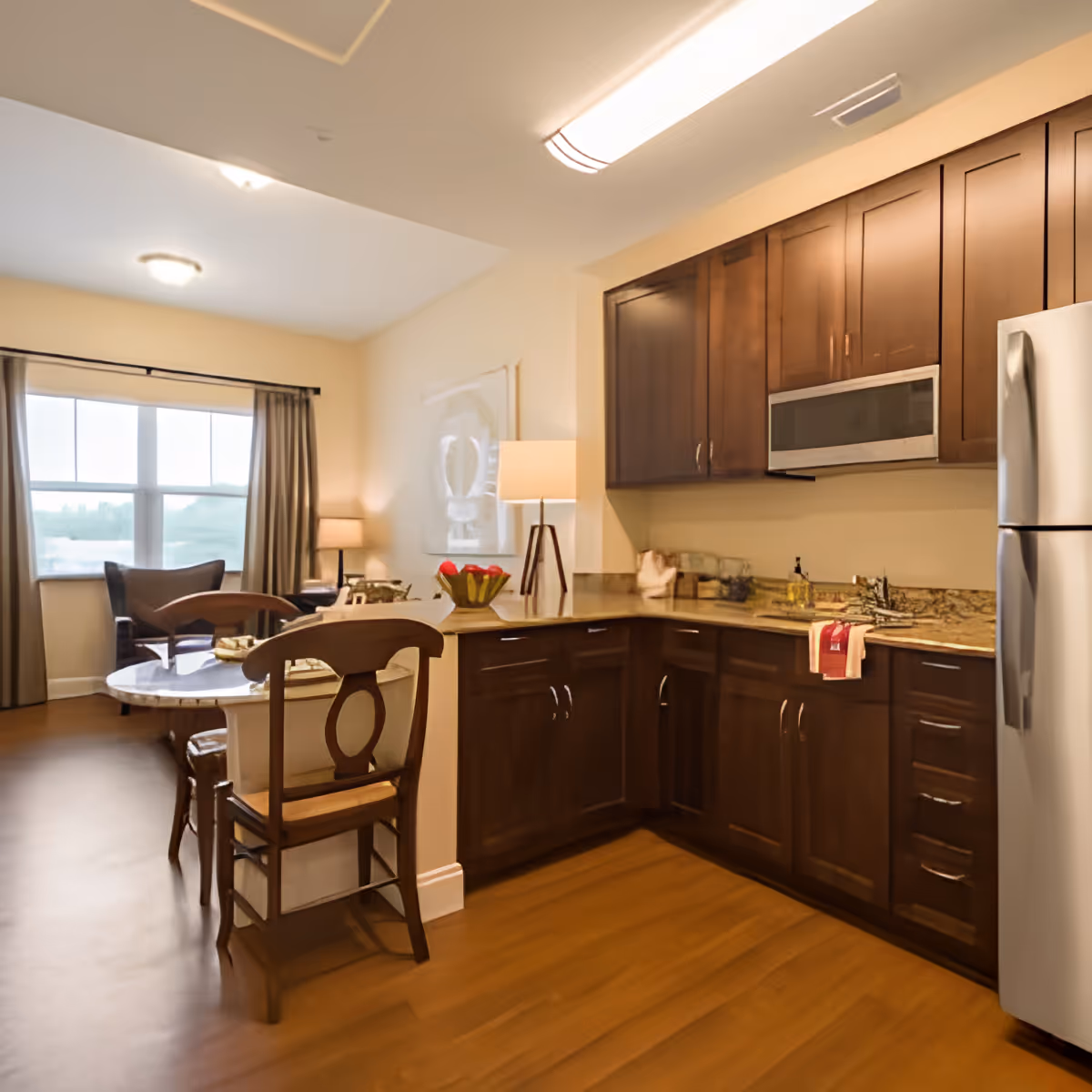 A cozy kitchen area with dark wooden cabinets, a stainless steel refrigerator, microwave, and granite countertops. Adjacent to the kitchen is a small dining area with a round table and wooden chairs. In the background, there is a living space with a large window, curtains, and an armchair, illuminated by soft lighting from lamps.