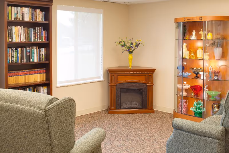 A cozy living room corner with two upholstered armchairs facing a wooden electric fireplace. To the left is a tall bookshelf filled with books, and to the right is a glass display cabinet with various decorative glass items. A window with a white blind is on the wall behind the fireplace, which has a yellow vase with flowers on top.
