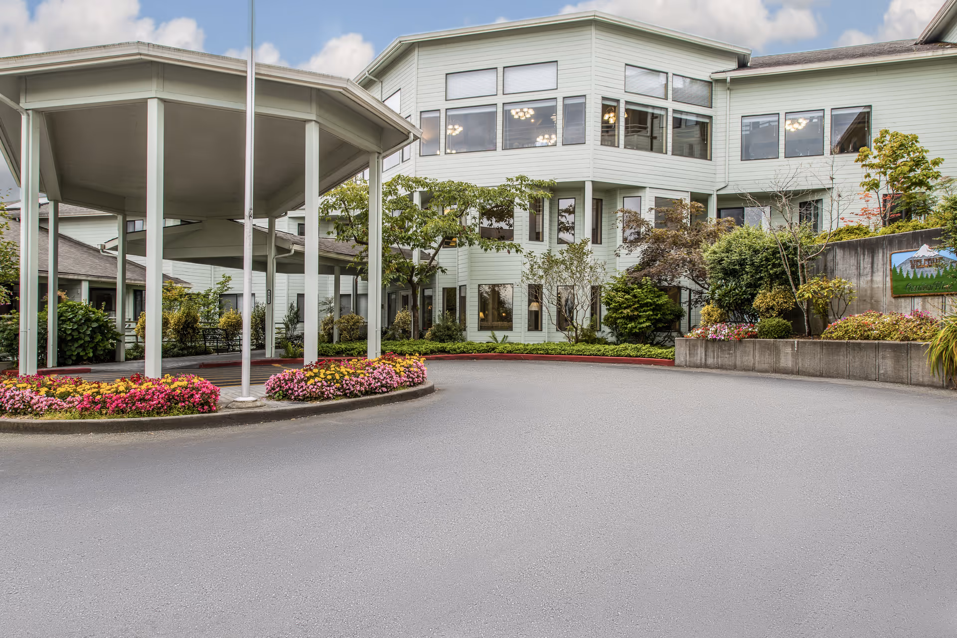 Front driveway and covered drop-off entrance of a multi-story senior living building with landscaped flower beds and shrubs.