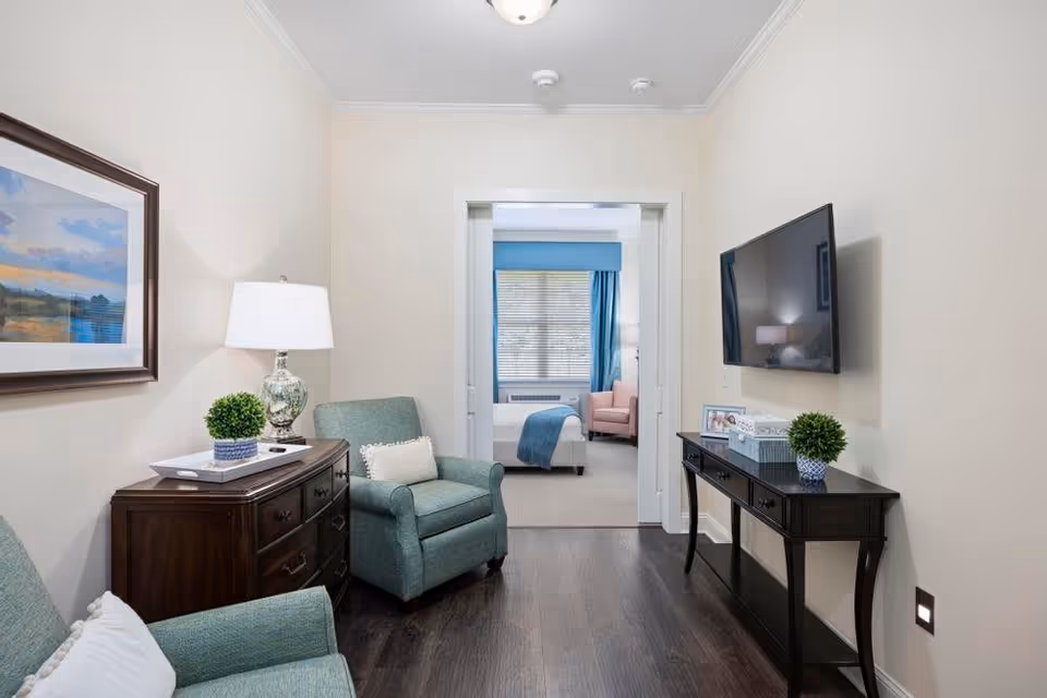 A cozy living area with two teal armchairs, a wooden side table with a lamp and a small plant, and a black console table with a plant and tissue box. A flat-screen TV is mounted on the wall. Through an open doorway, a bedroom with a bed, a pink armchair, and a window with blue curtains is visible.