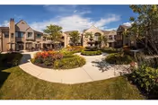 Outdoor courtyard area of a senior living community with a central fountain surrounded by landscaped flower beds, pathways, and two-story buildings under a blue sky with some clouds.