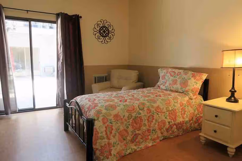 Bedroom with a floral-patterned single bed, nightstand and lamp, armchair, and sliding glass door to the outside.