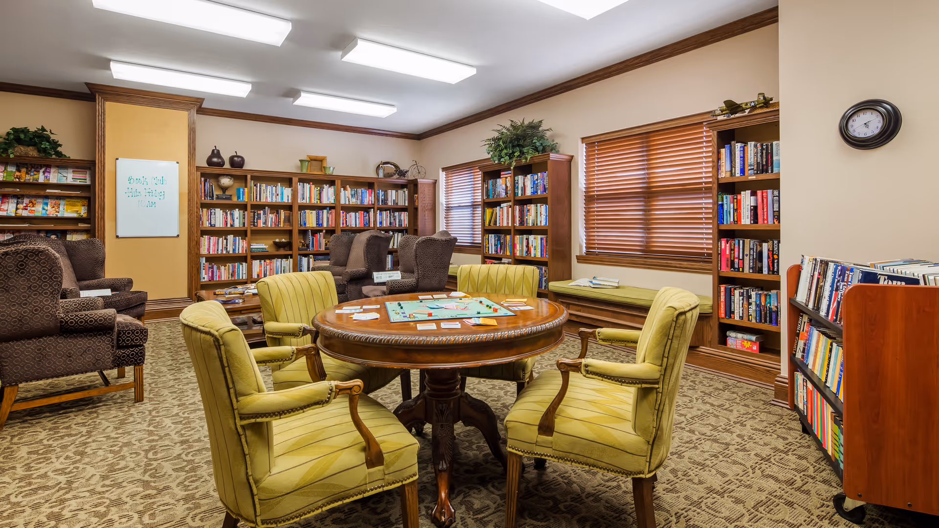 A cozy library room with wooden bookshelves filled with books, a round wooden table with a board game set up, and six green upholstered chairs around the table. There are additional brown patterned armchairs in the background, a window with wooden blinds, a clock on the wall, and a whiteboard with a note about a book club meeting.