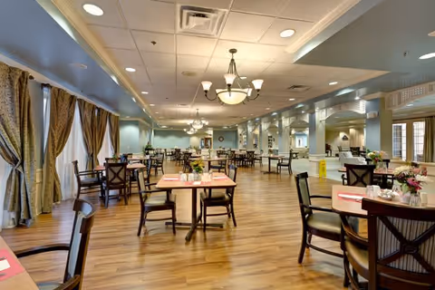 Spacious dining room with wooden floors, multiple tables and chairs arranged neatly, large windows with curtains allowing natural light, and elegant ceiling lights providing warm illumination.