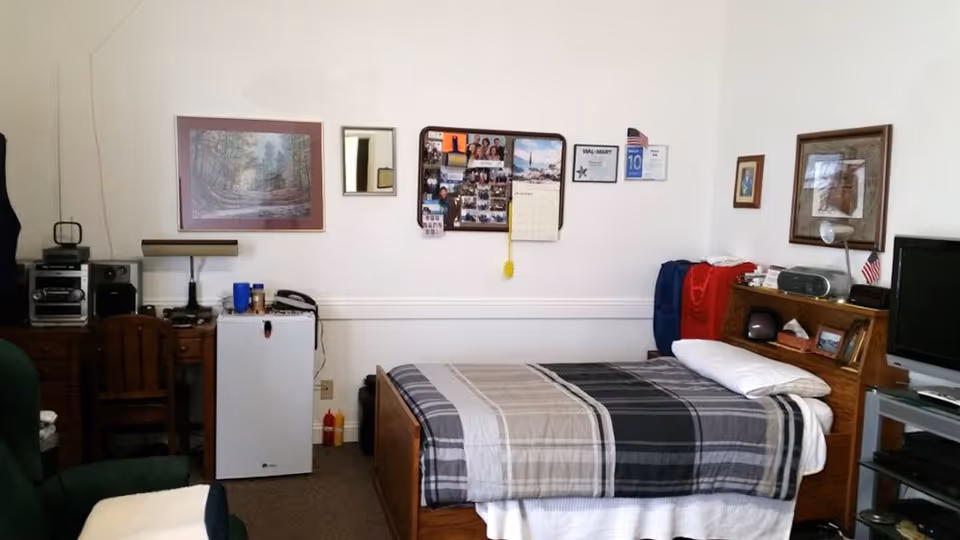 A small, tidy bedroom in a senior living facility with a single bed covered in a plaid blanket. The room includes a wooden desk with a chair, a small white mini-fridge, a green armchair, and a TV on a stand. The walls are decorated with framed pictures, a calendar, and a bulletin board with photos and notes.