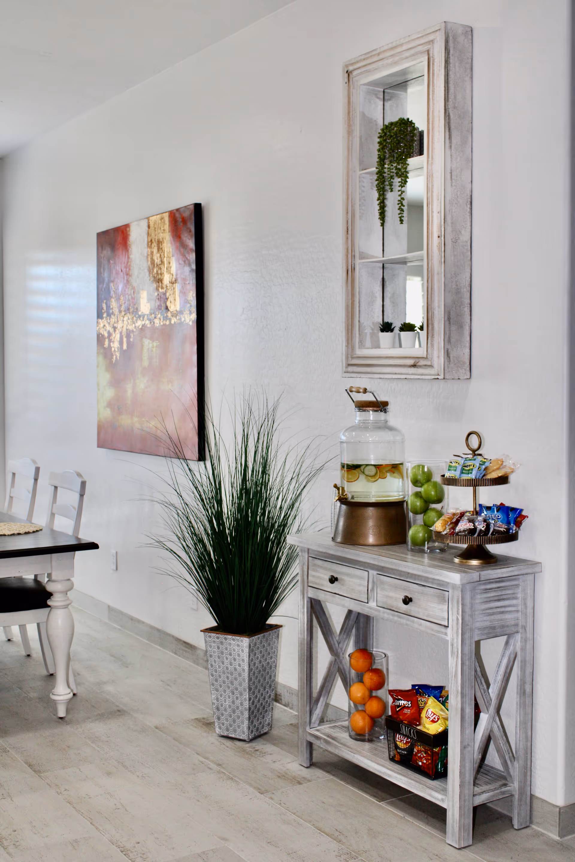 A bright interior space featuring a white wooden console table with drawers. On the table, there is a glass beverage dispenser filled with lemon water, a glass container with green apples, and a two-tiered tray holding various snacks. Below the table, there is a glass container with oranges and several bags of chips. To the left of the table, a tall potted plant with long green leaves stands on the floor. A colorful abstract painting hangs on the white wall above a dining table with white chairs partially visible on the left side. A small wall-mounted cabinet with glass panes and small potted plants inside is above the console table.