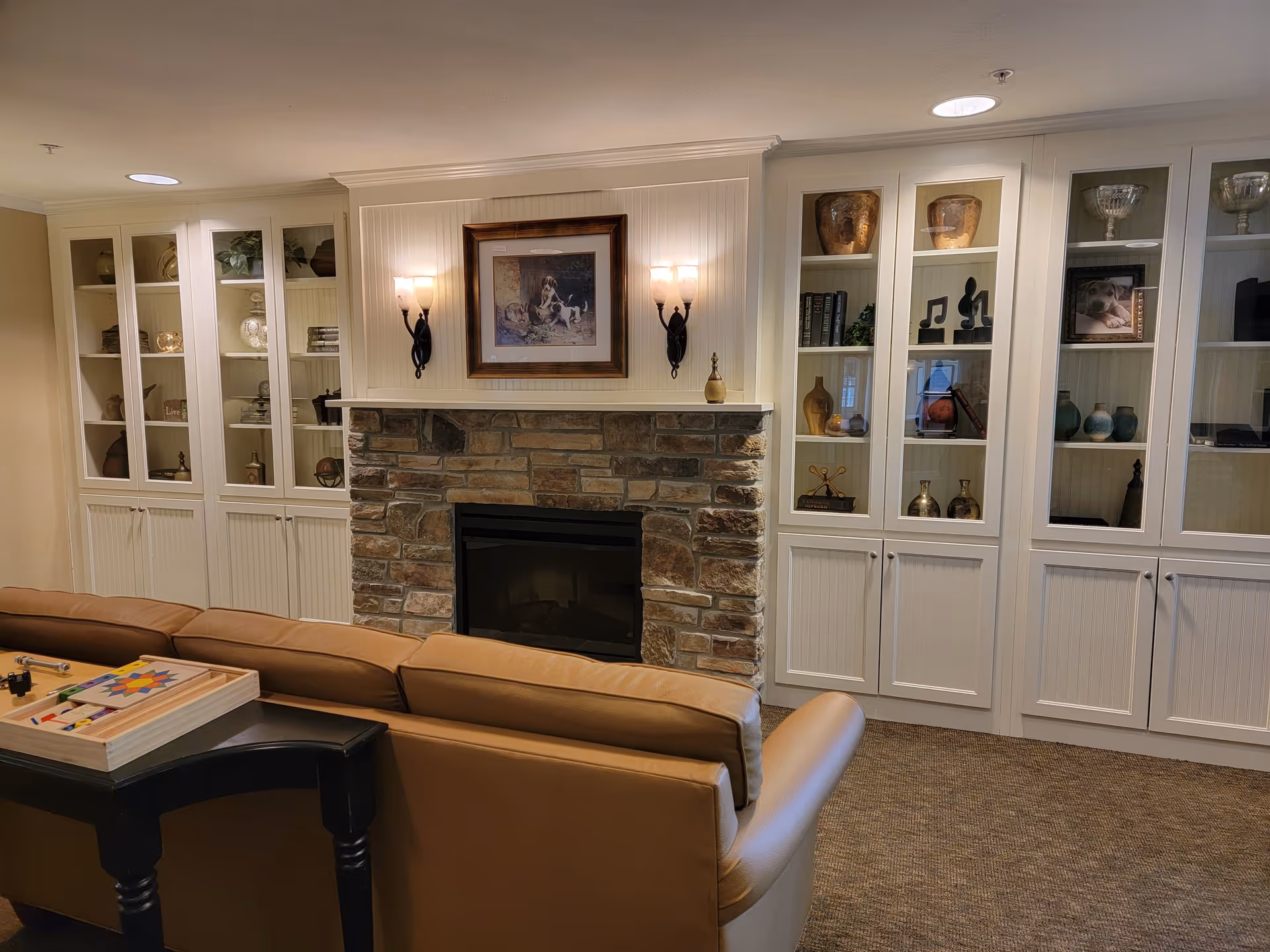 A cozy living room area with a stone fireplace in the center, flanked by white built-in cabinets filled with decorative items and books. Above the fireplace is a framed picture of dogs, and two wall sconces with lights are mounted on either side. In front of the fireplace is a brown leather sofa with a small black table holding a wooden board game.