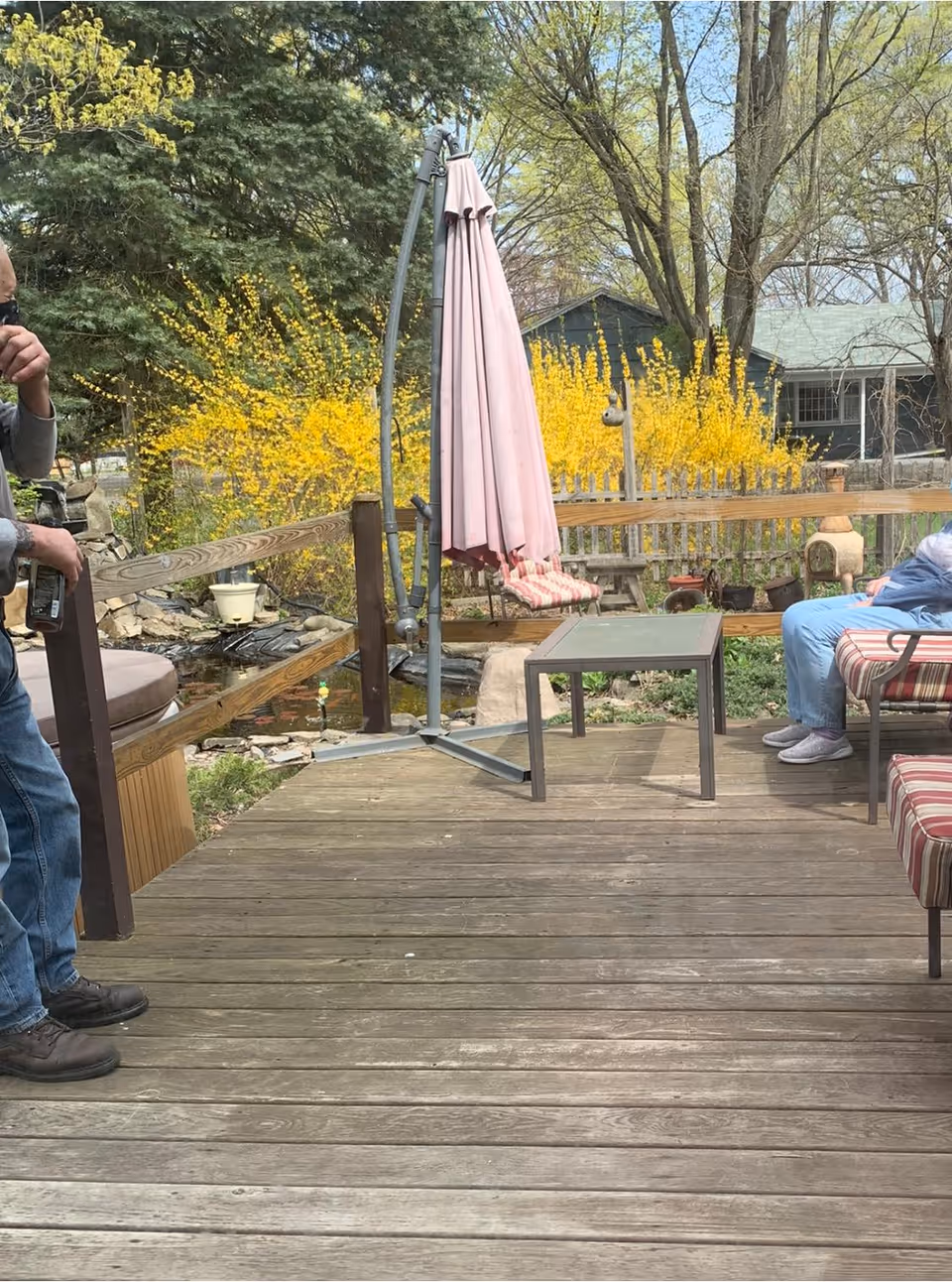 Outdoor wooden deck area with a folded pink patio umbrella, a small table, and striped cushioned chairs. There are yellow flowering bushes and trees in the background, along with a wooden fence and a small pond. Two people are partially visible, one standing on the left and one sitting on the right side of the deck.