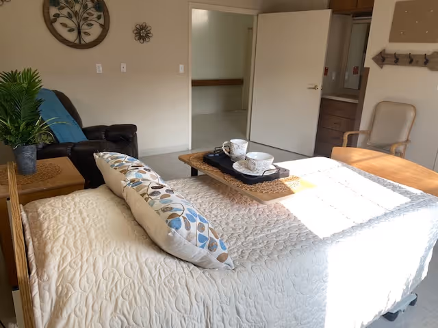 A cozy bedroom with a bed covered in a white quilt and decorative pillows. A wooden tray with two cups and saucers sits on the bed. To the left, there is a small table with a potted plant and a dark recliner chair with a blue blanket. On the wall above the chair, there are two decorative wall hangings. An open door reveals a hallway and part of a wooden chair is visible on the right side of the image.