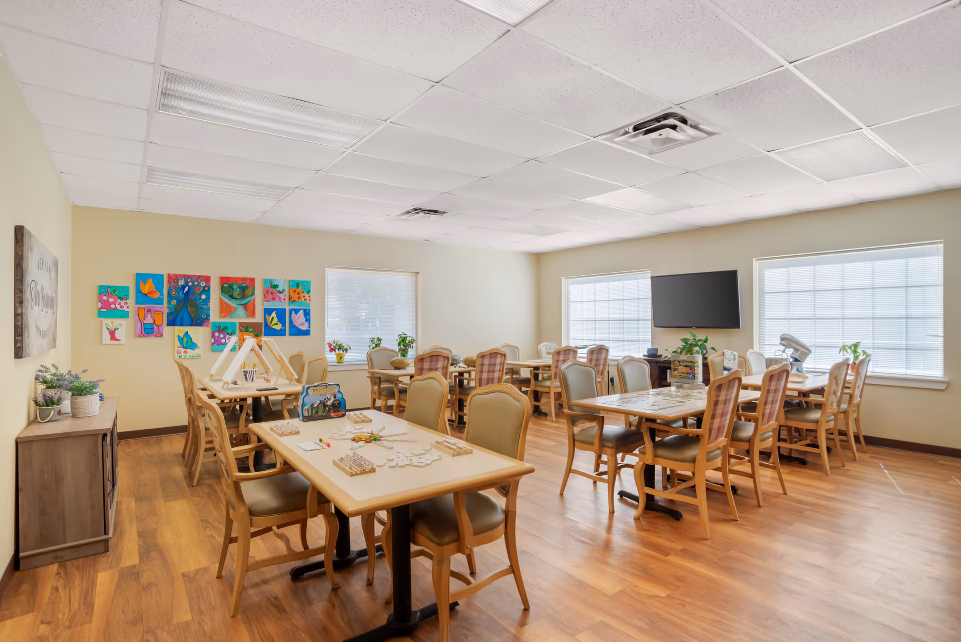 A bright activity room with several tables and chairs arranged for group activities. The tables have puzzles and games on them. The walls are decorated with colorful artwork and there are large windows letting in natural light. A flat-screen TV is mounted on one wall, and there are plants on the windowsills and a side cabinet.