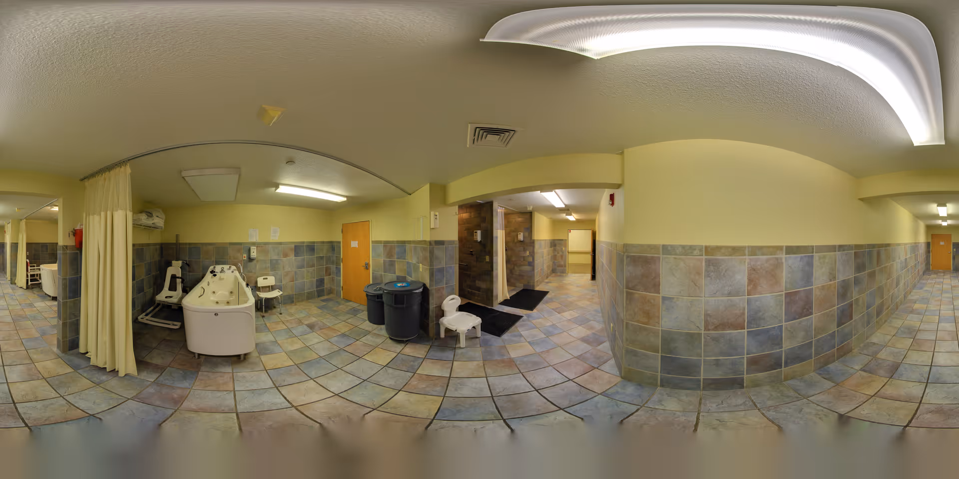 A tiled room with a large bathtub designed for assisted bathing, a shower chair, and a privacy curtain. The walls are partially tiled with multicolored stone tiles, and the ceiling has fluorescent lighting. There are two trash bins and a small white chair near the bathtub. The room appears to be part of a senior living facility's bathing area.