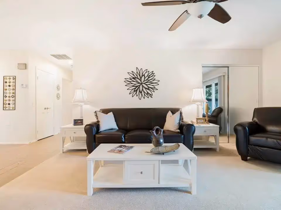 A bright living room with a black leather sofa and matching armchair. The sofa has two white pillows. There is a white coffee table in front of the sofa with decorative items on it. Two white side tables with lamps flank the sofa. A decorative metal wall art piece hangs above the sofa. A ceiling fan with dark wooden blades is visible on the white ceiling. A doorway with mirrored sliding doors is on the right side, reflecting part of the room and a window with a view of greenery outside.