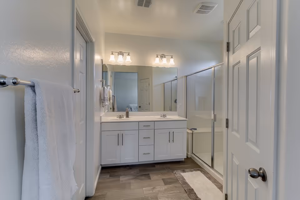 A clean, modern bathroom with a double sink vanity featuring white cabinets and silver handles. Above the sinks are two sets of three-light fixtures and a large mirror. To the right is a glass-enclosed shower with a sliding door. A white towel hangs on a silver towel rack on the left wall, and a white and brown bath mat is placed on the wooden floor in front of the shower.