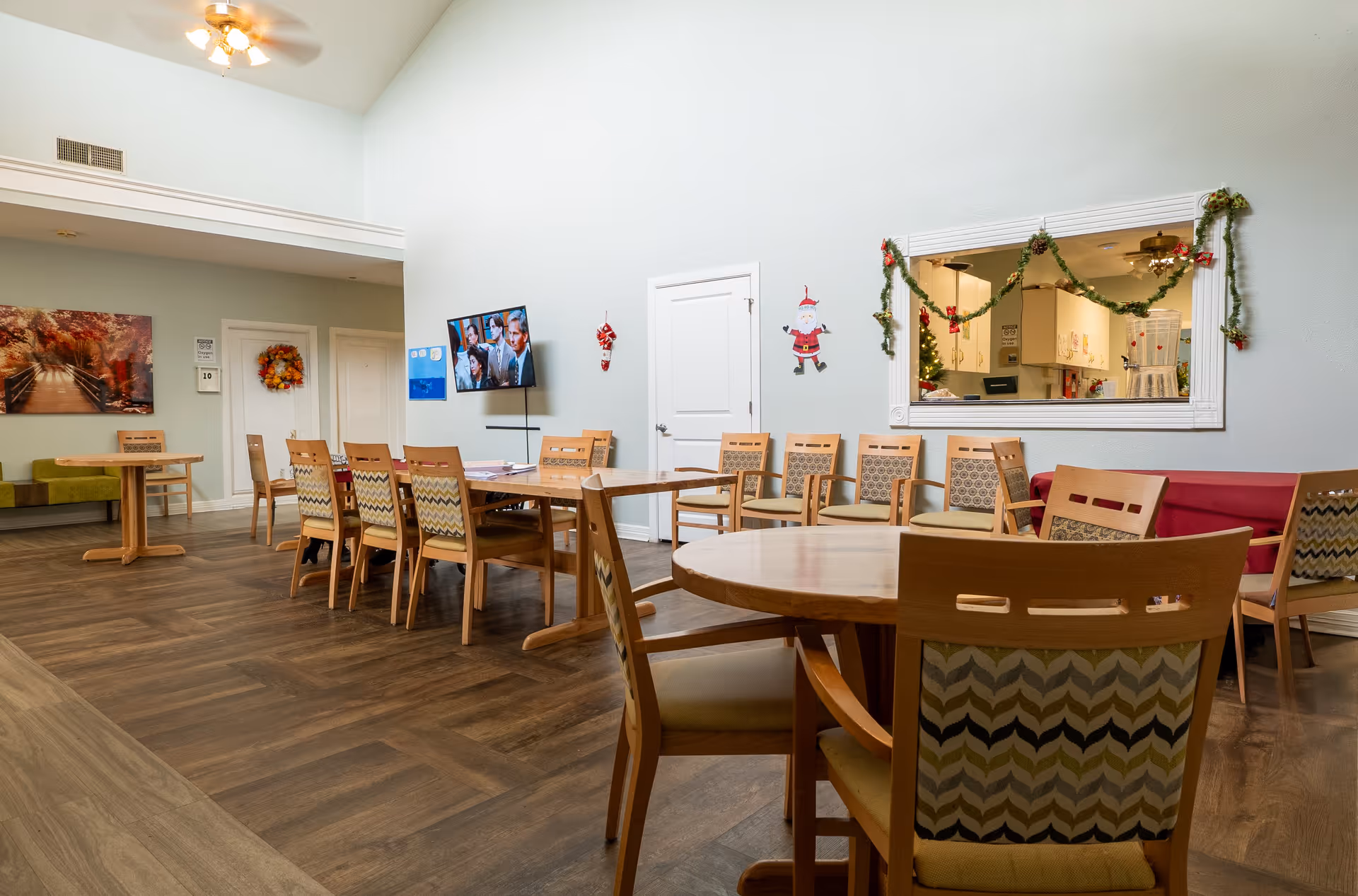 A senior living facility dining room with multiple wooden tables and chairs arranged neatly. The room has a high ceiling with a ceiling fan, a wall-mounted TV showing a scene from a show or movie, and holiday decorations including garlands and a Santa Claus figure on the wall. There is a large window opening to a kitchen area decorated with Christmas garlands.