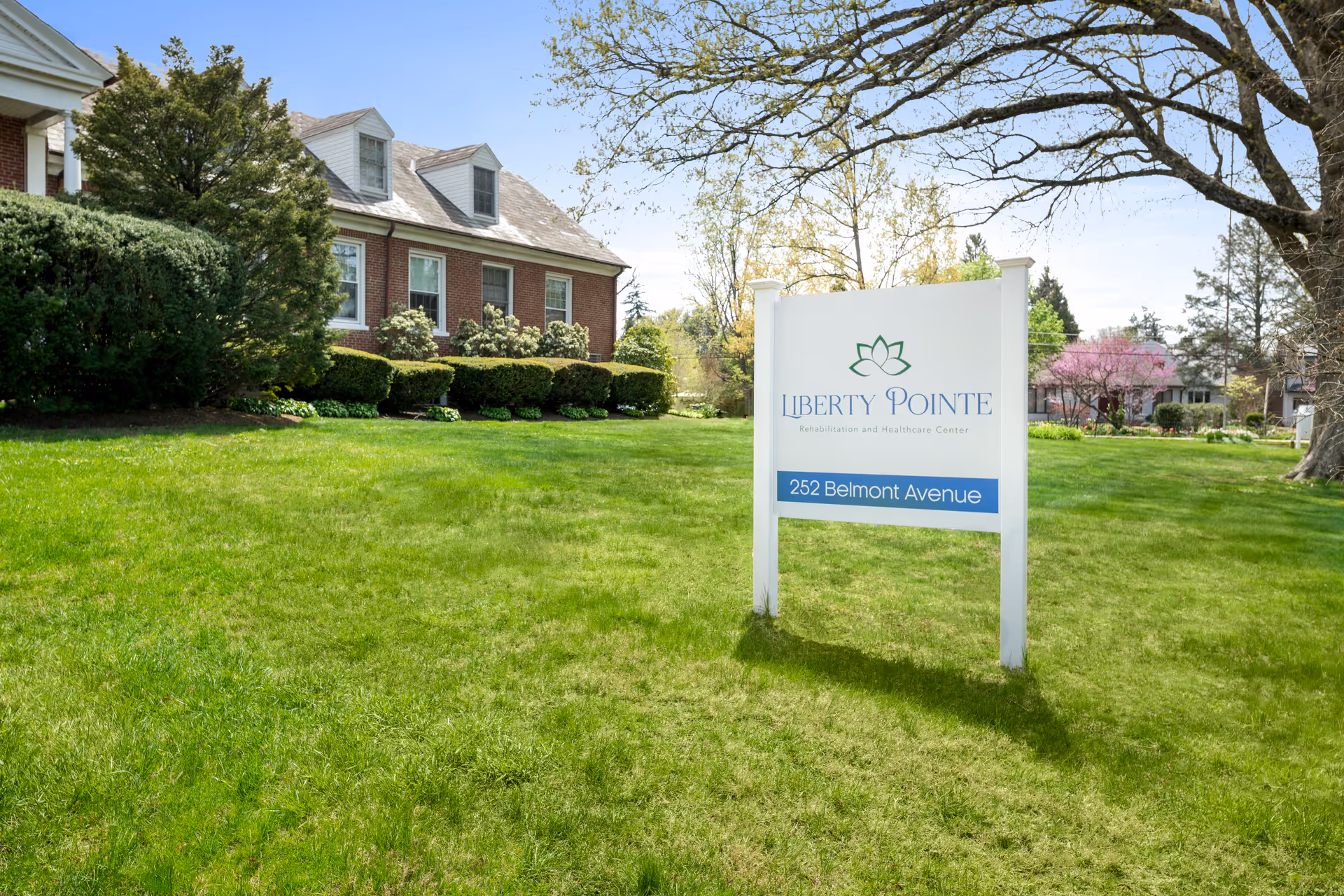 A white sign reading 'Liberty Pointe Rehabilitation and Healthcare Center' stands on a green lawn in front of a brick building and trees.