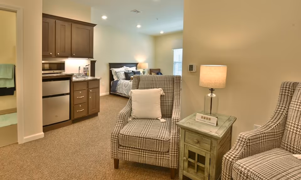 A cozy assisted living apartment featuring a small kitchenette with dark wood cabinets, a mini fridge, and a microwave. In the foreground, there are two patterned armchairs with a small wooden side table and a lamp between them. In the background, a bed with pillows and a bedside lamp is visible, along with a window letting in natural light. To the left, a bathroom entrance with towels is partially visible.