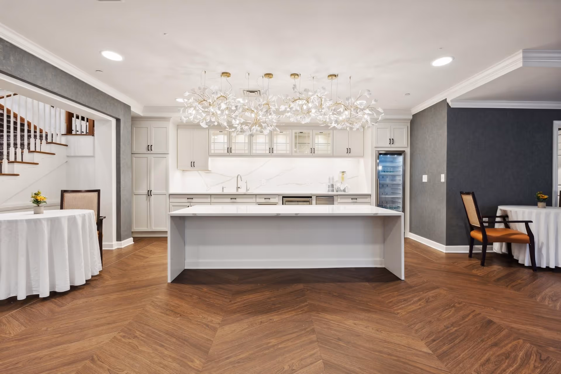 A modern kitchen area with a large white island countertop in the center, white cabinetry with glass doors, a built-in refrigerator, and elegant pendant lighting featuring floral designs. The floor has a wood pattern, and there are two round tables with white tablecloths and chairs on either side of the kitchen space. A staircase with white railings is visible on the left side.