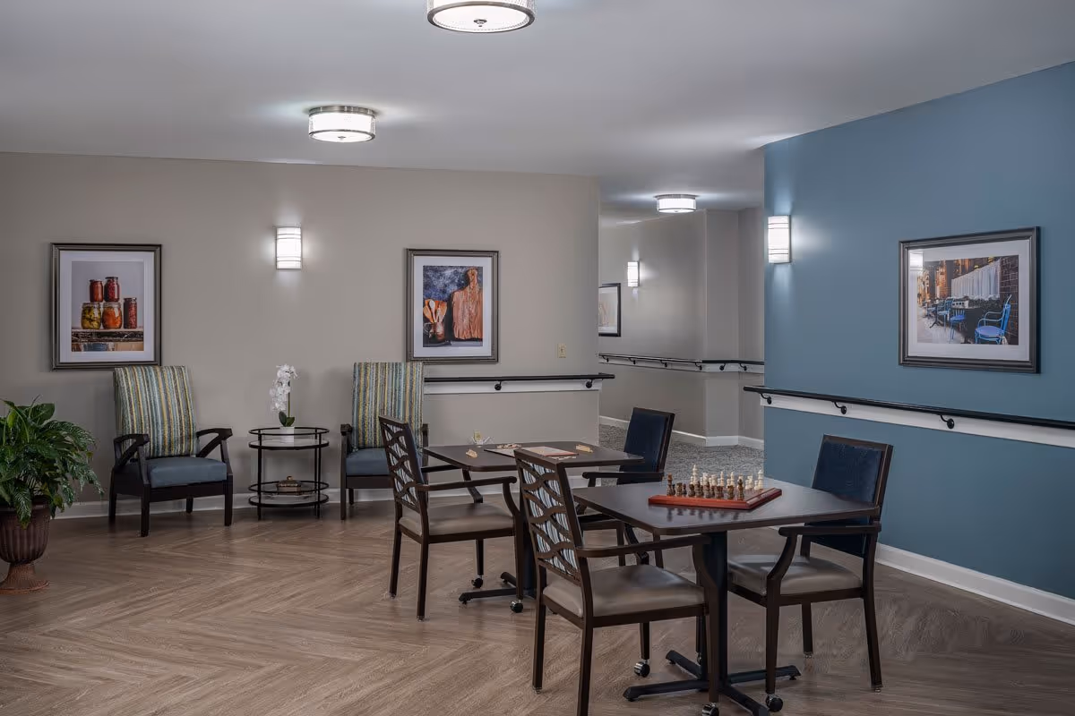 A well-lit common area with two tables and chairs arranged for playing board games, including chess and checkers. The room features light wood flooring, a blue accent wall with framed artwork, and two striped armchairs with a small round table between them. There are ceiling and wall lights, handrails along the walls, and a potted plant in the corner.