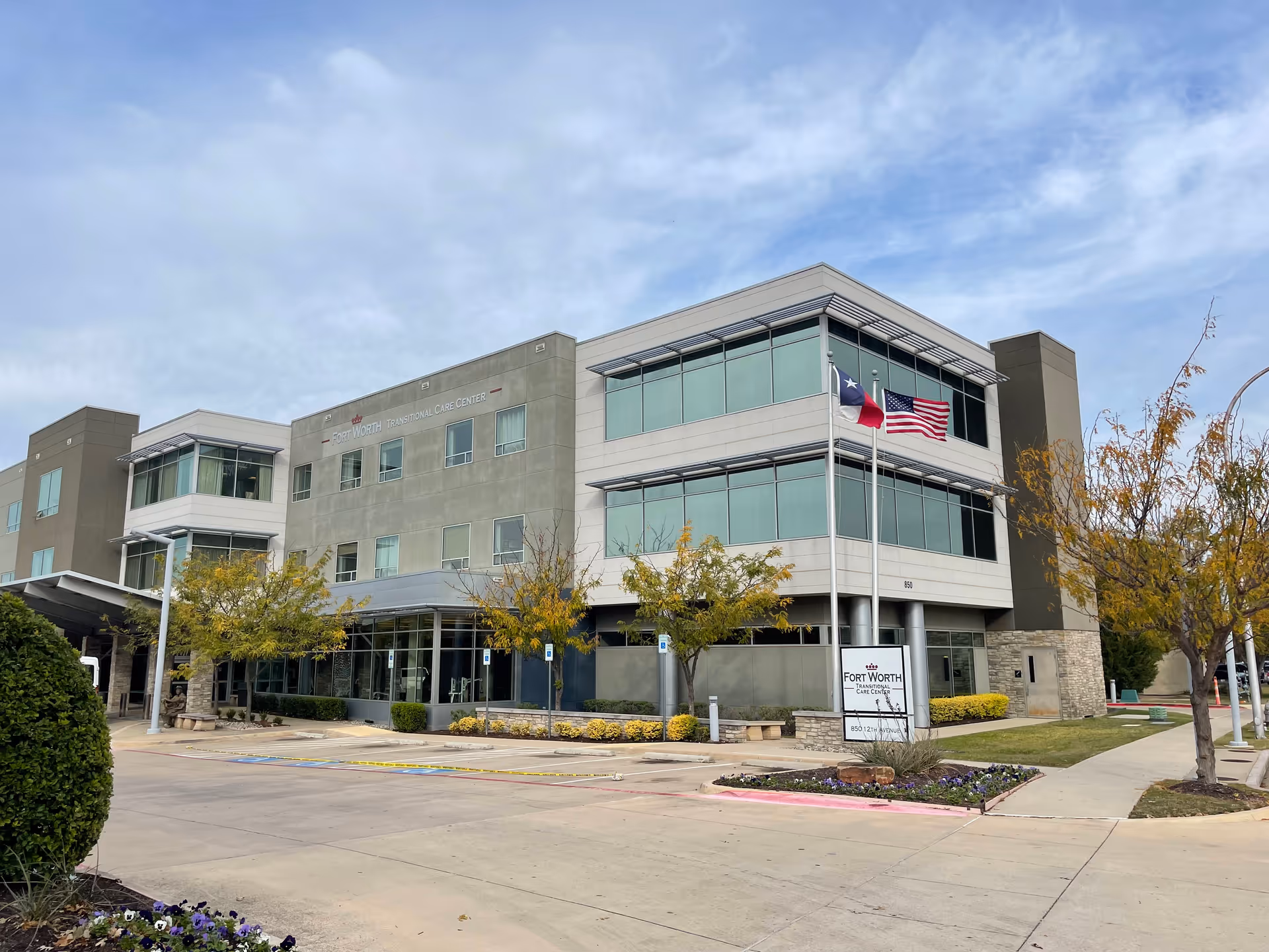 Exterior view of the Fort Worth Transitional Care Center building, a modern three-story structure with large windows, two flagpoles displaying the Texas and United States flags, landscaped trees and bushes, and a clear sky above.