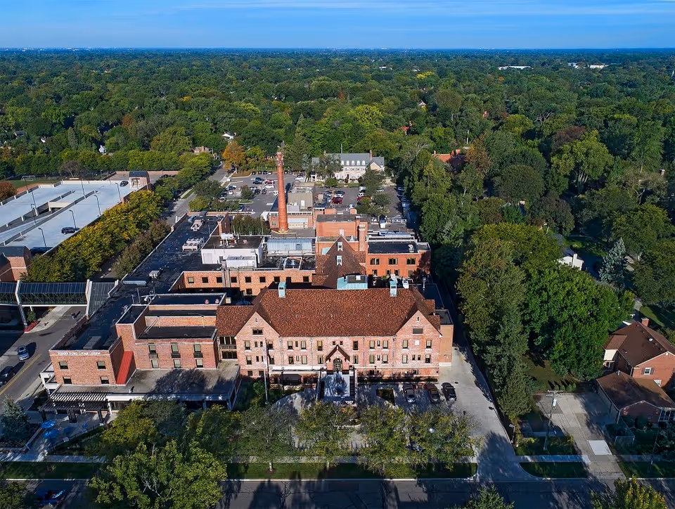Aerial view of a large brick building complex surrounded by trees and residential houses, with a parking lot and a tall chimney visible on the property under a clear blue sky.