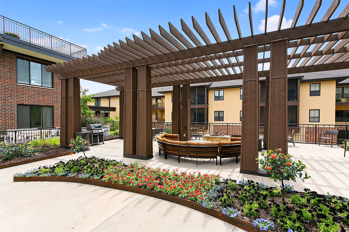 Outdoor patio area at Folkestone facility featuring a large wooden pergola with a curved seating arrangement around a fire pit. There are flower beds with colorful flowers in the foreground, a barbecue grill to the left, and multi-story residential buildings in the background under a partly cloudy sky.