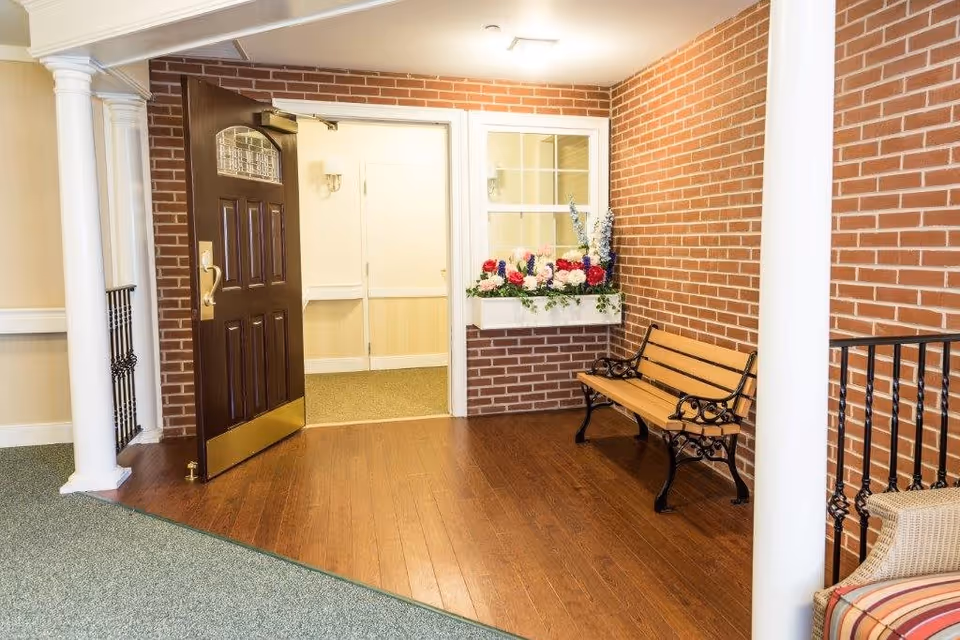 Interior view of a senior living facility entrance area with a dark wooden door open to a hallway. There is a wooden bench with black metal armrests and legs against a brick wall. A window box filled with colorful flowers is mounted on the brick wall above the bench. The floor is a combination of wood and carpet, and white columns and railings are visible.