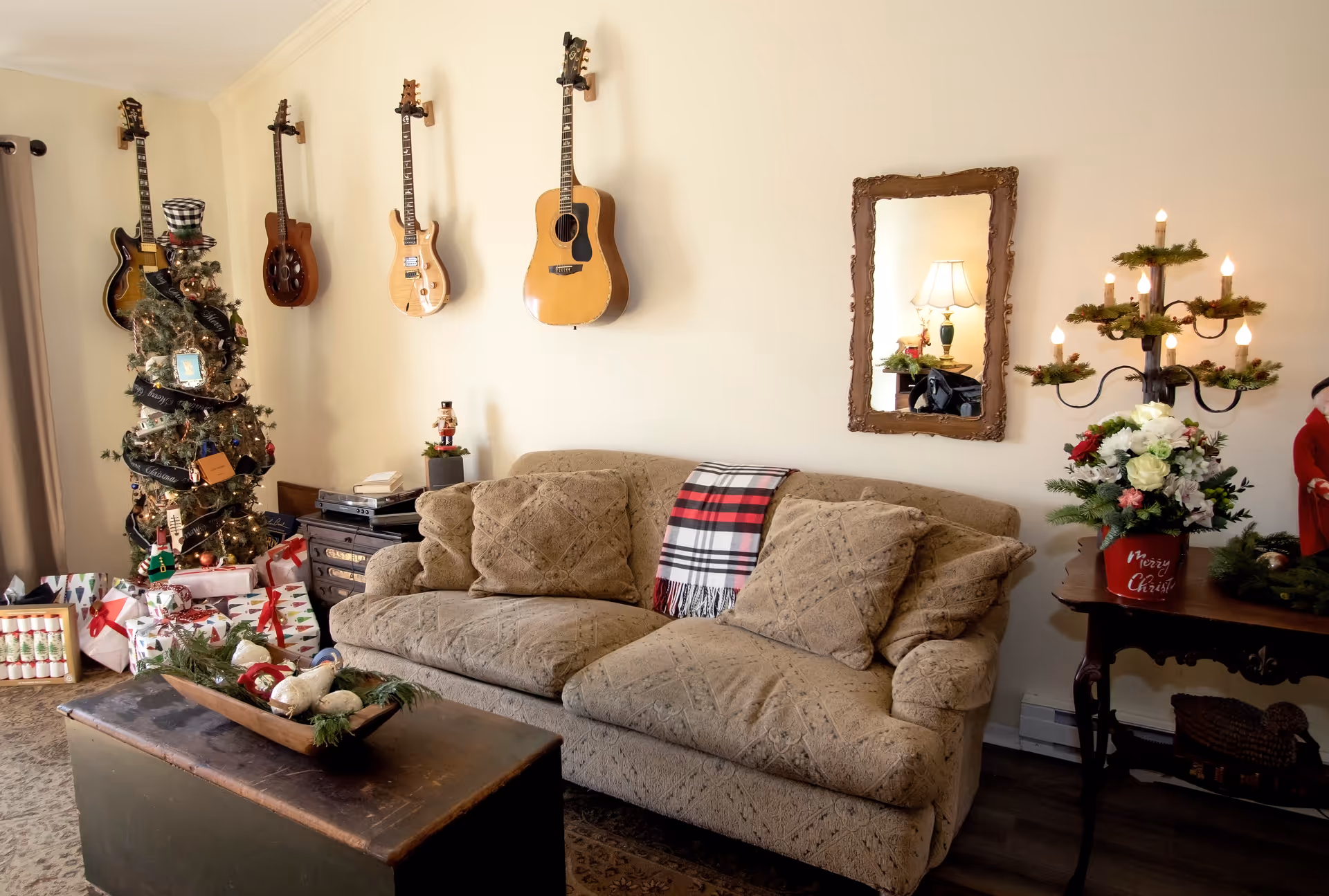 A cozy living room decorated for Christmas with a beige patterned sofa, a plaid throw blanket, and a wooden coffee table with holiday decorations. Four guitars are mounted on the wall above the sofa. A Christmas tree with presents underneath is in the corner, and a decorative candelabra with lit candles and flowers sits on a side table next to a framed mirror reflecting a lamp.
