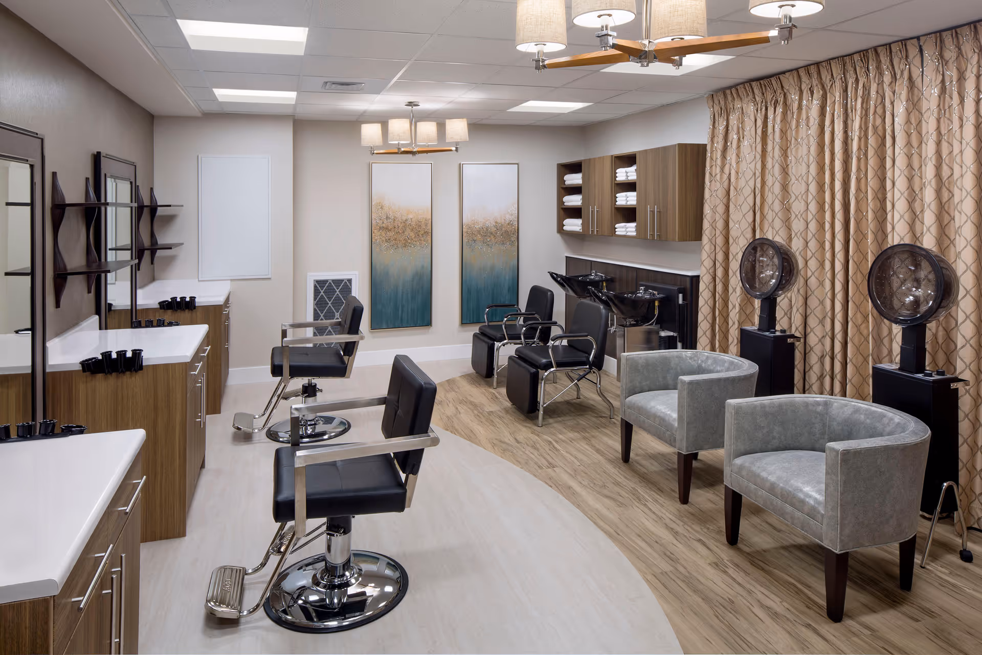 A modern salon area in a senior living facility featuring black salon chairs in front of mirrors and sinks for hair washing. The room has wood and white cabinetry, two gray armchairs, two hair dryers, beige patterned curtains, and two abstract paintings on the wall.