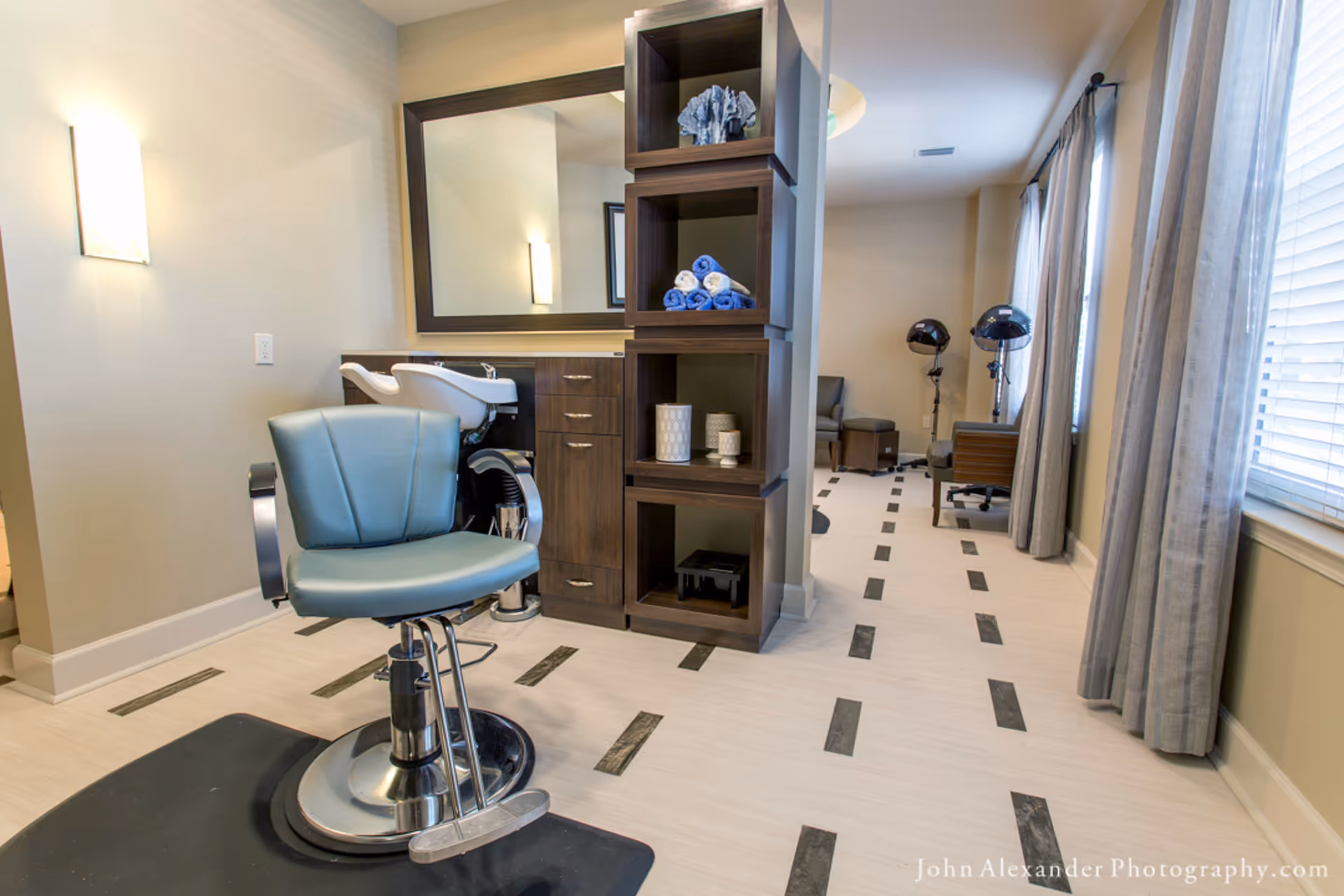 A salon-style interior with a styling chair and sink in front of a mirror, shelving with towels, and hooded hair dryers by a window.