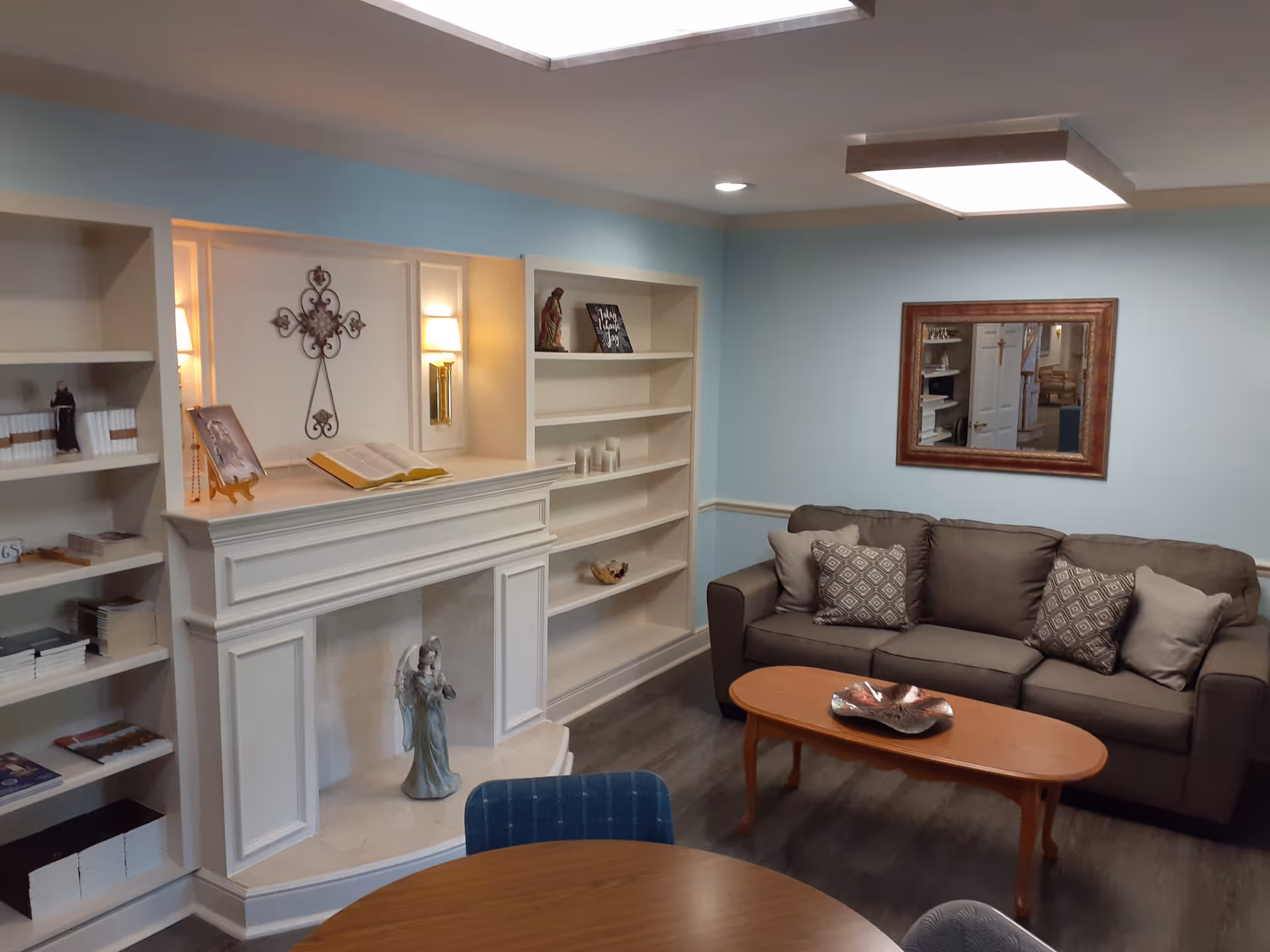 A cozy living room with a brown sofa adorned with patterned and plain cushions, a wooden coffee table with a decorative bowl, built-in white shelves with various decorative items and books, a white fireplace with a statue and an open book on the mantel, soft wall lighting, a large mirror on the light blue wall, and a wooden round table with blue chairs in the foreground.