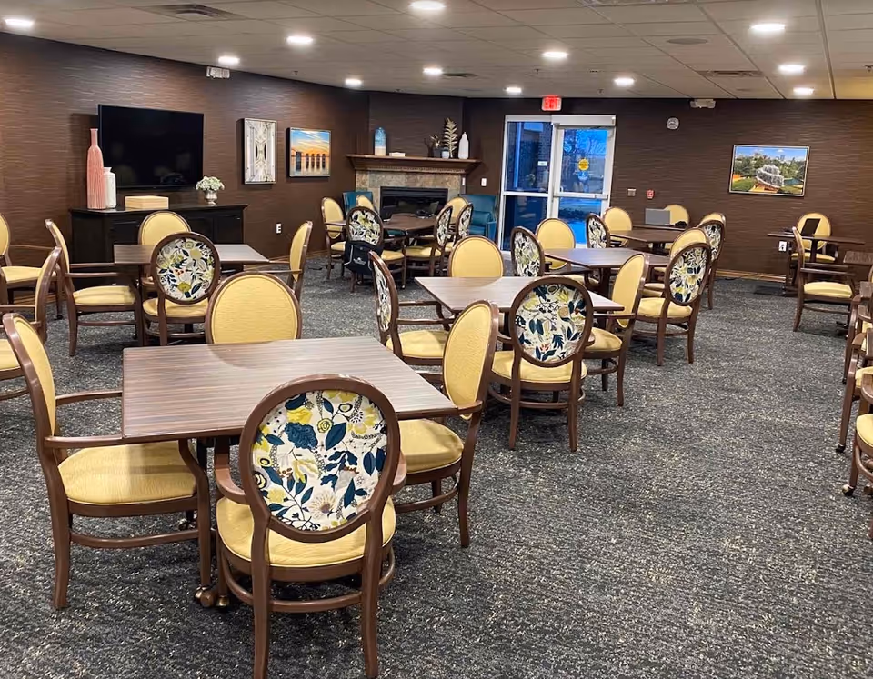 A spacious dining room with multiple wooden tables and chairs featuring yellow cushions and floral-patterned backs. The room has a carpeted floor, brown walls adorned with framed artwork, a flat-screen TV mounted on one wall, and a fireplace with decorative items on the mantel. There is a glass door at the back leading outside.