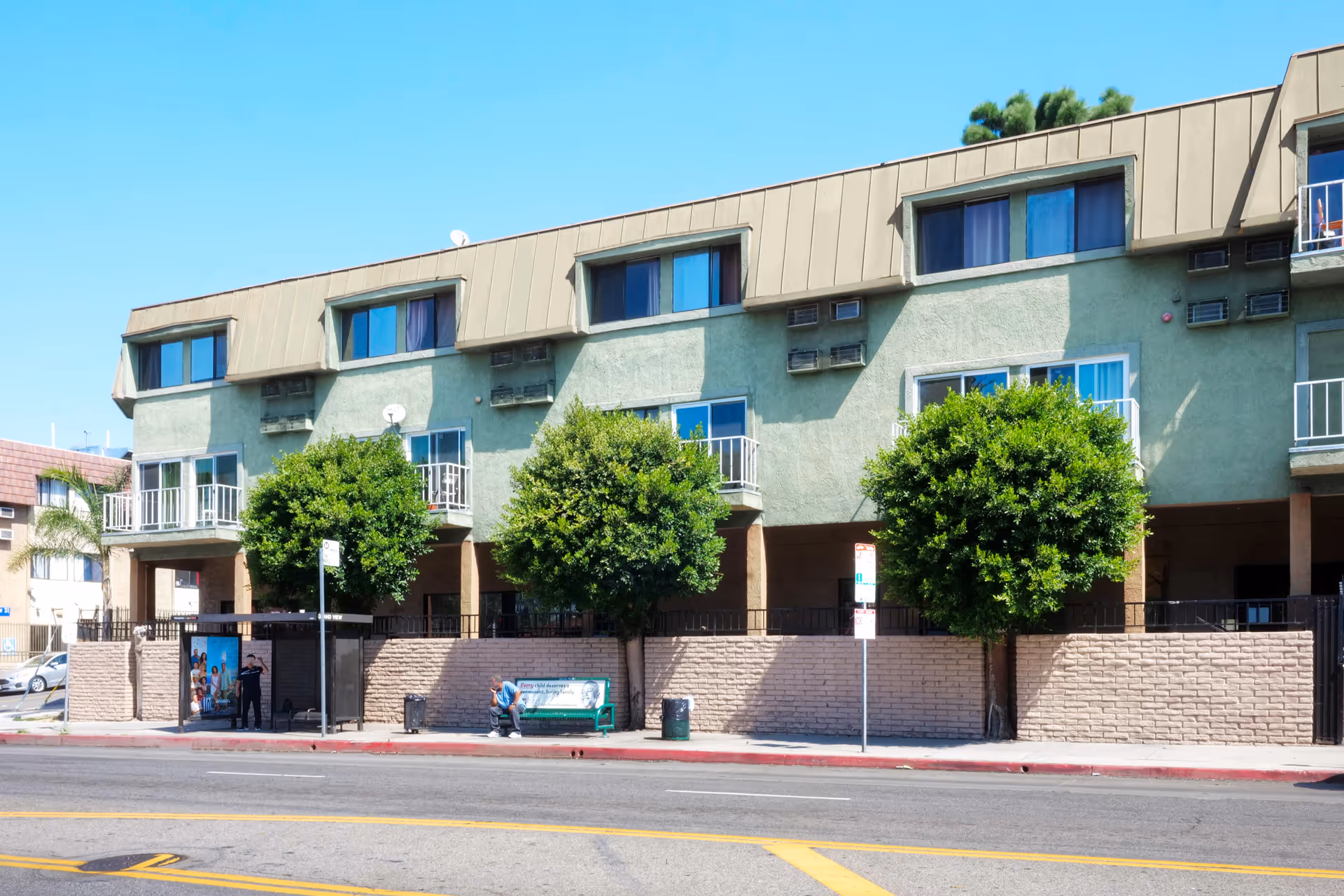 Exterior view of a multi-story residential building with green walls and beige roof panels. The building has several windows and small balconies. In front of the building, there is a sidewalk with a bus stop shelter, a green bench, three neatly trimmed trees, and a brick wall. A person is sitting on the bench, and another person is standing near the bus stop. The sky is clear and blue.