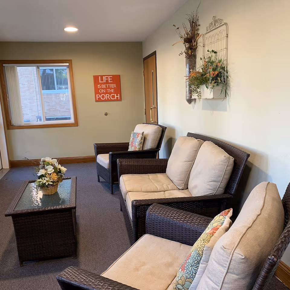 A cozy indoor seating area with three cushioned wicker chairs and a glass-top wicker coffee table with a floral arrangement. On the wall, there is a decorative piece with plants and a sign that reads 'LIFE IS BETTER ON THE PORCH'. A window and a door are visible in the background.