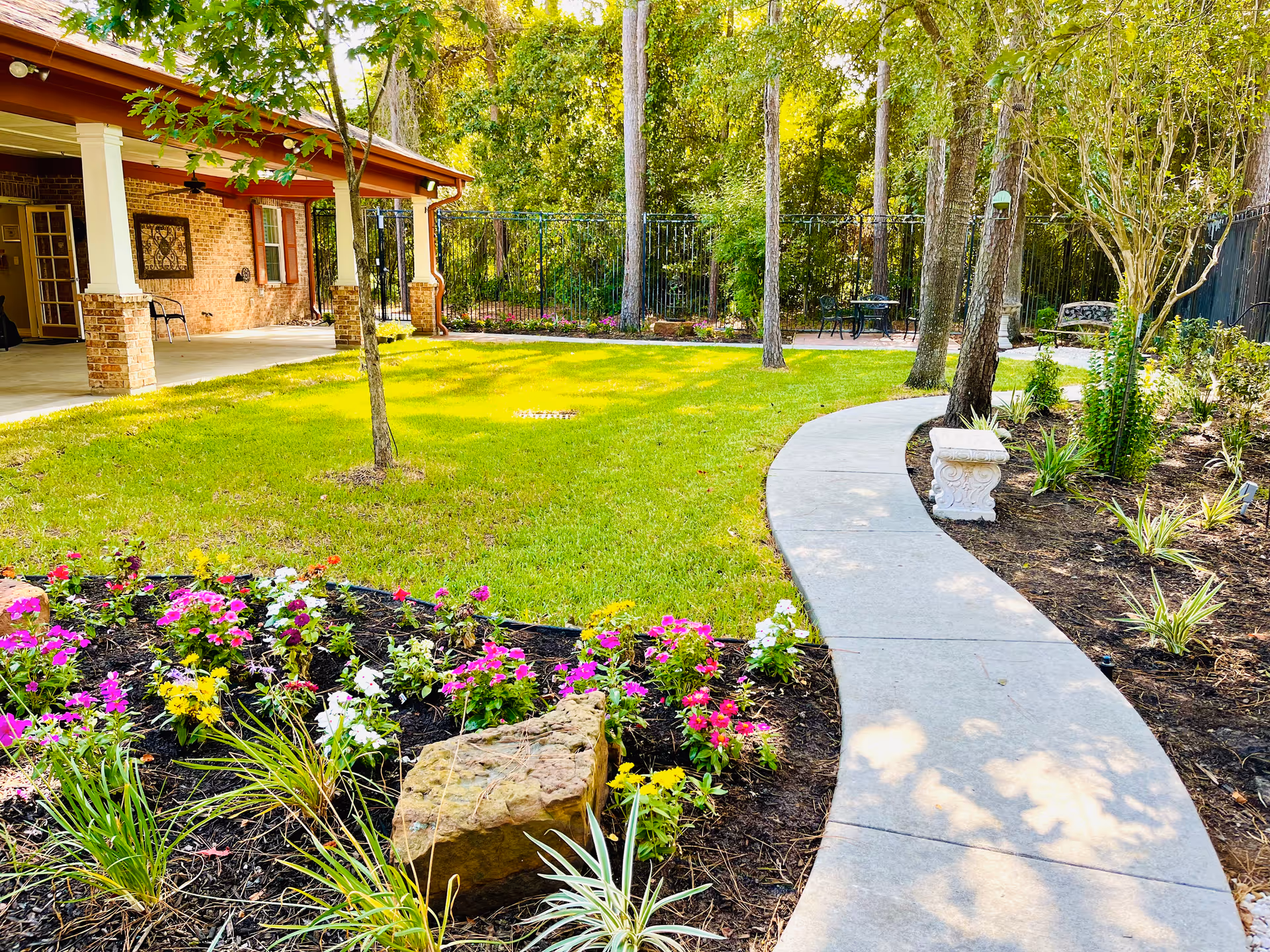Curved concrete path through a landscaped yard with colorful flower beds, trees, and a covered brick patio.