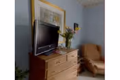 Dresser topped with a television and a vase of flowers beside an armchair under framed artwork in a pastel blue room.
