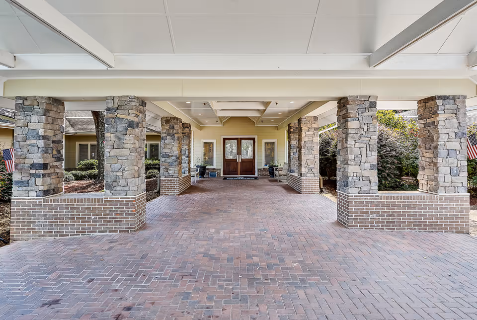 Covered entrance area of a building with stone pillars and brick flooring leading to double wooden doors. There are benches and plants near the entrance, and American flags are visible on the sides.